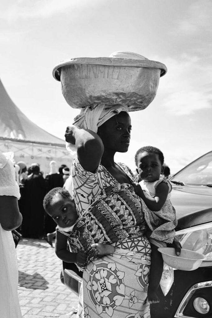 A Ghanaian mother carrying two children with a basin on her head, Nalerigu market scene.
